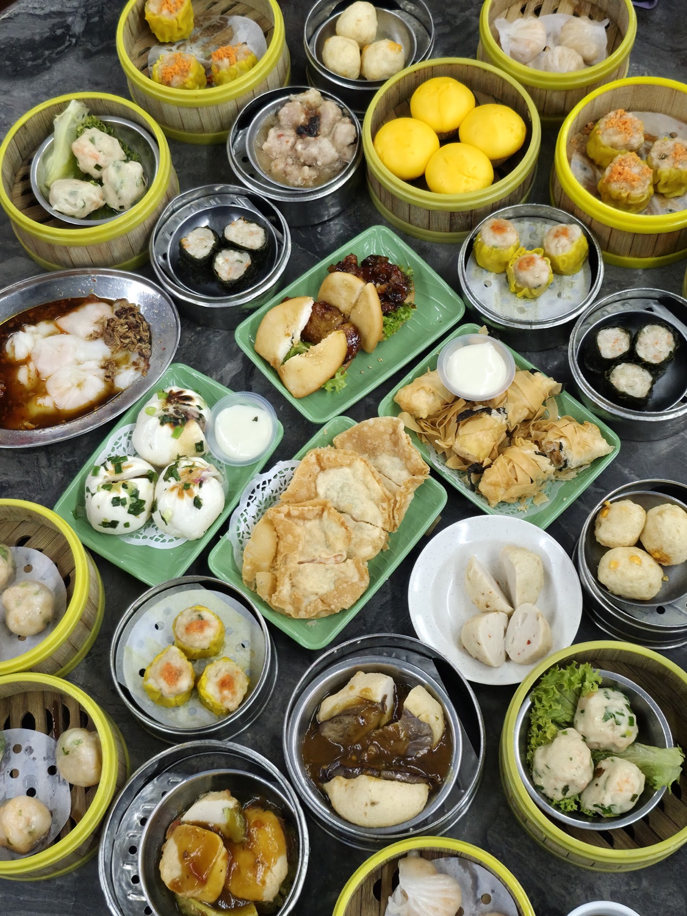 A spread of steamed dim sum baskets — har gao, siu mai, char siu bao, custard buns and lo mai gai — set out across a tea-stained table at Maxim